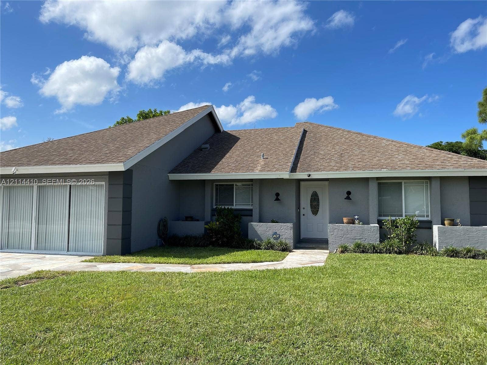 Single-story exterior featuring a gray stucco facade, a multi-gabled roofline, stone-tiled front walkway, and attached multi-panel sliding glass doors