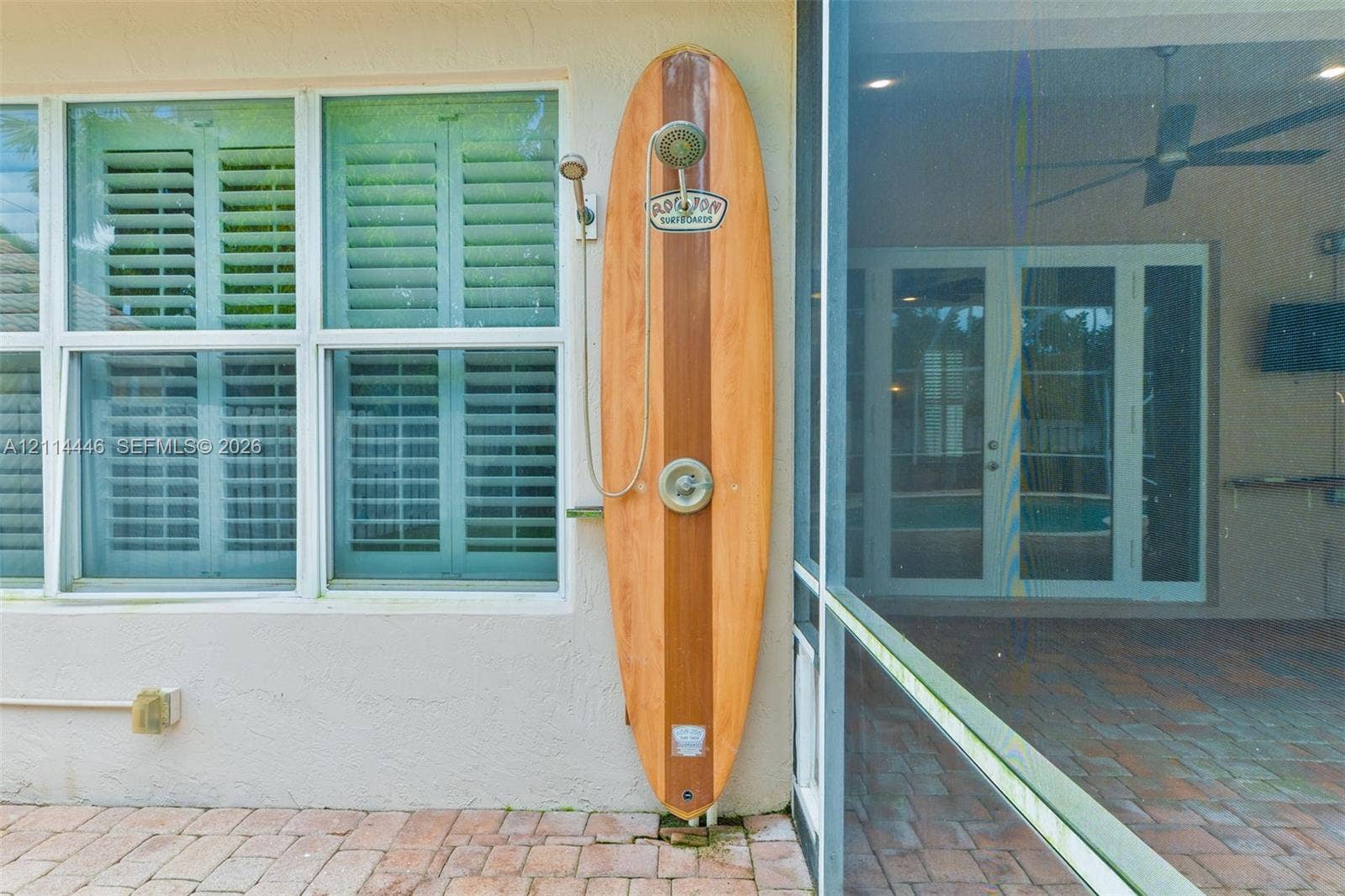 Outdoor shower featuring a wood-finished surfboard mounting, polished metal shower head, and wall-mounted faucet, situated on a brick-paver patio with exterior plantation shutters