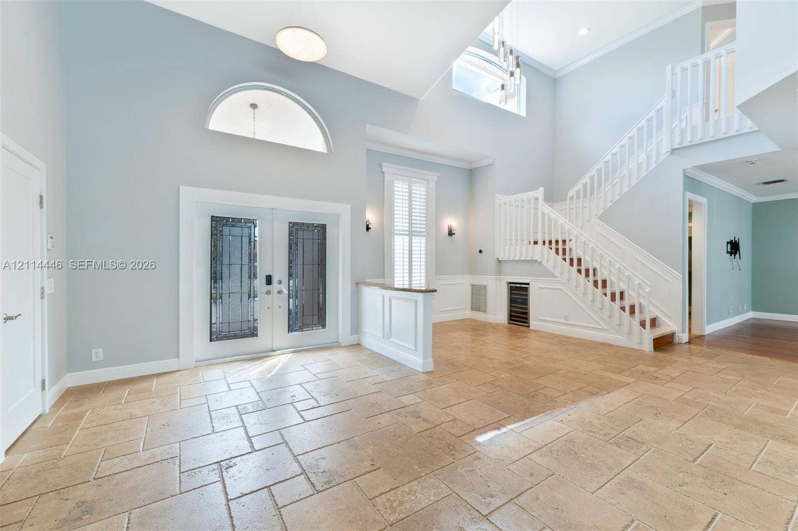 Double-height entryway featuring large-format tile flooring, a grand staircase with decorative balustrades, double entry doors with glass inserts, an arched transom window, and built-in under-stair cabinetry