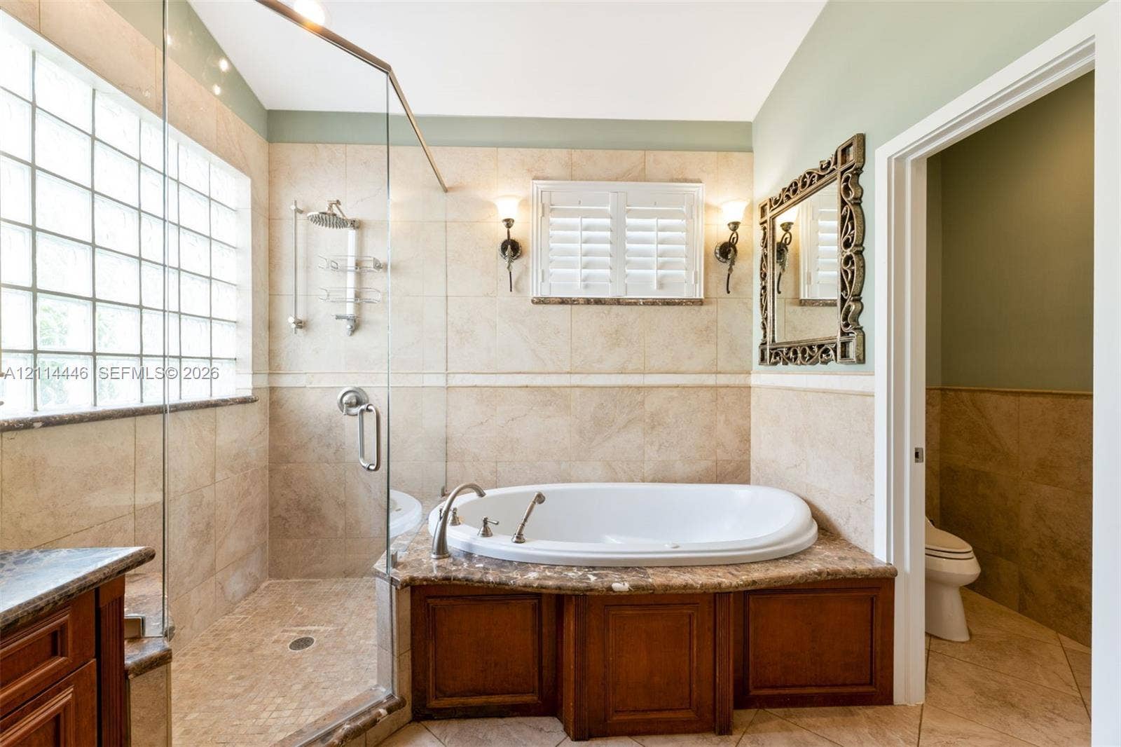 Walk-in shower with glass enclosure, jetted soaking tub with stone-surface surround, wood-finish cabinetry, and glass block window