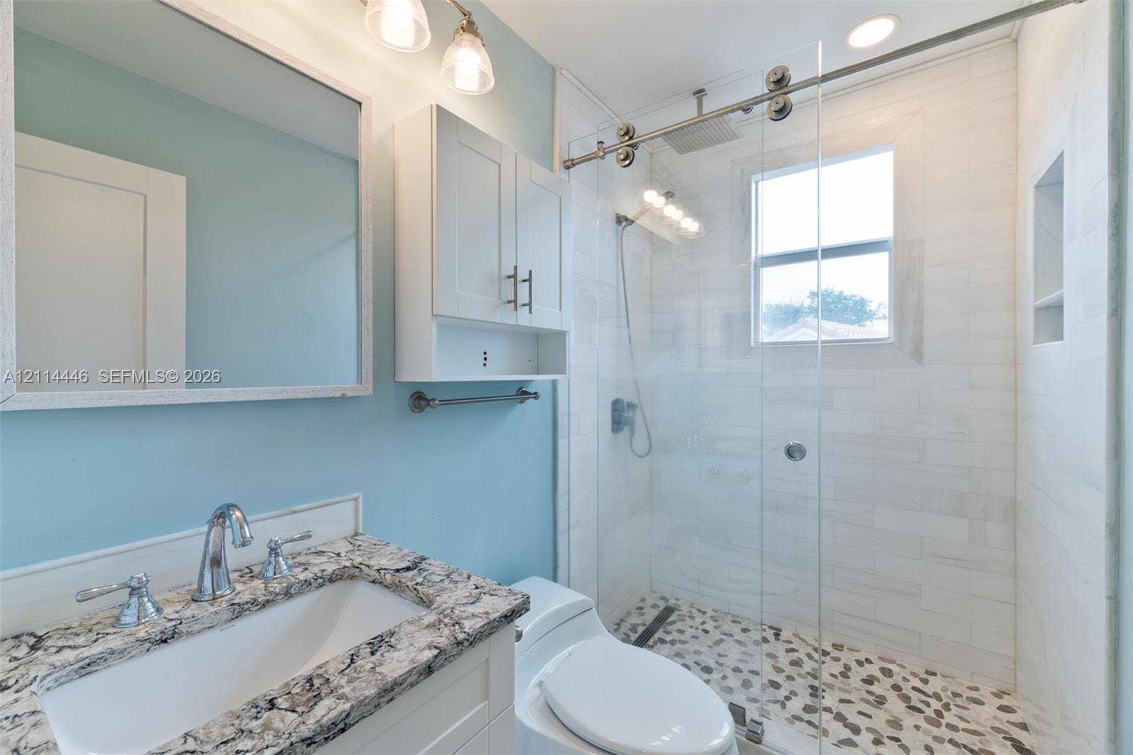 Bathroom featuring a stone-top vanity with an undermount sink, walk-in shower with a sliding glass door, pebble tile shower floor, and built-in recessed wall shelving