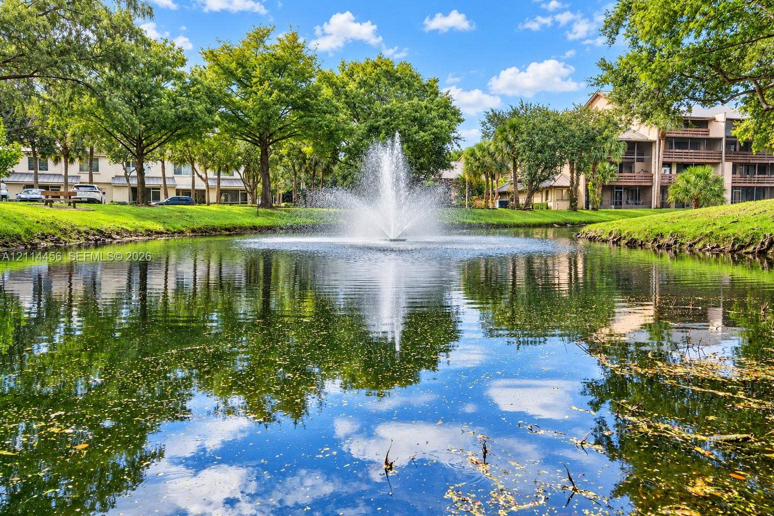 Water feature fountain centered within a pond, surrounded by mature trees and manicured lawns