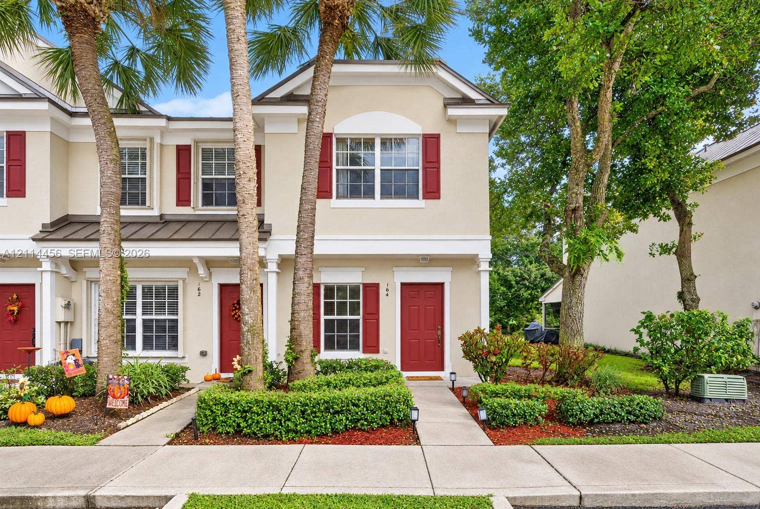 Two-story exterior featuring light-toned stucco, decorative shutters, a metal accent roof, and paved walkways surrounded by manicured landscaping and mature trees
