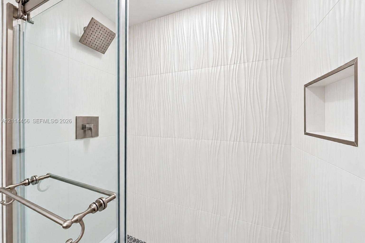 Walk-in shower featuring textured white wall tile, rain-style shower head, brushed metal fixtures, built-in recessed wall niche, and glass door enclosure