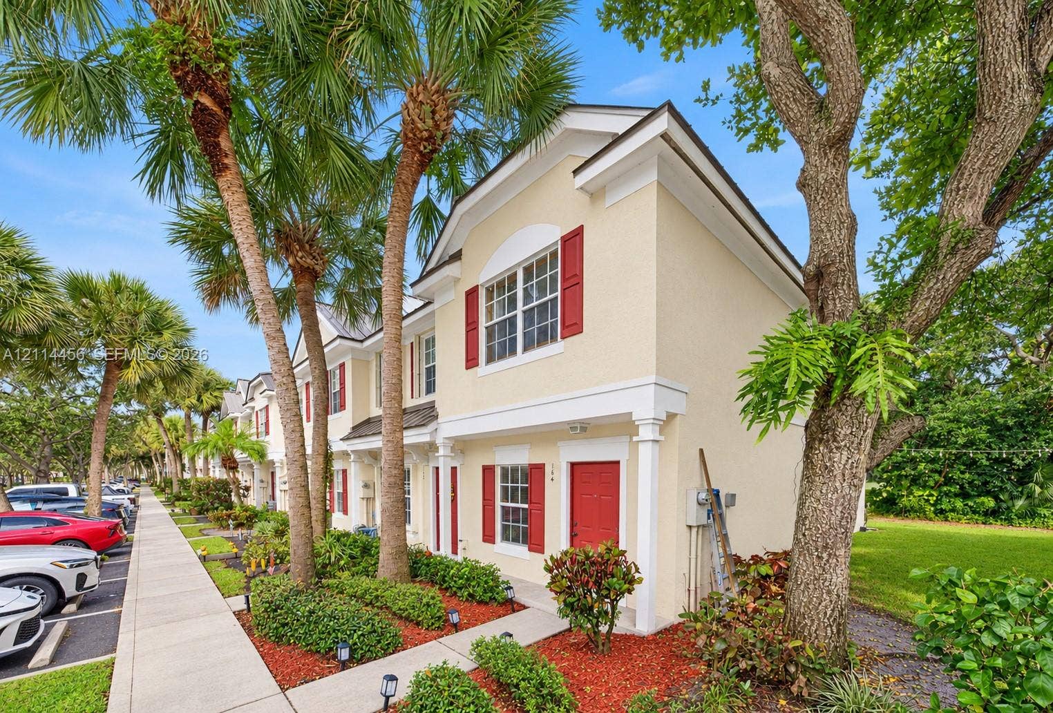 Two-story exterior featuring light-colored stucco siding, contrasting red shutters, decorative column accents, and mature palm tree landscaping