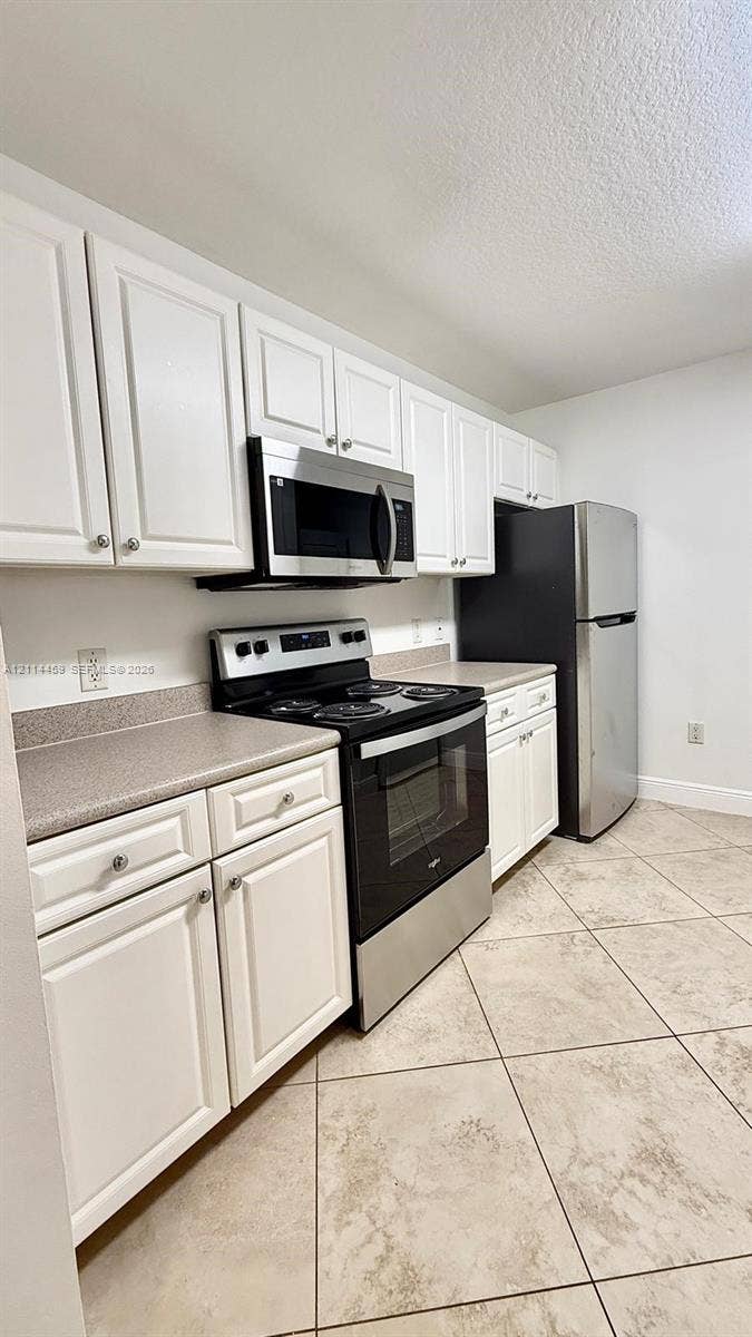 1203 Town Center Drive, #207, Jupiter, Jupiter, FL 33458 Kitchen featuring white shaker-style cabinetry, neutral solid-surface countertops, tile flooring, and stainless steel appliances including a built-in microwave, range, and refrigerator