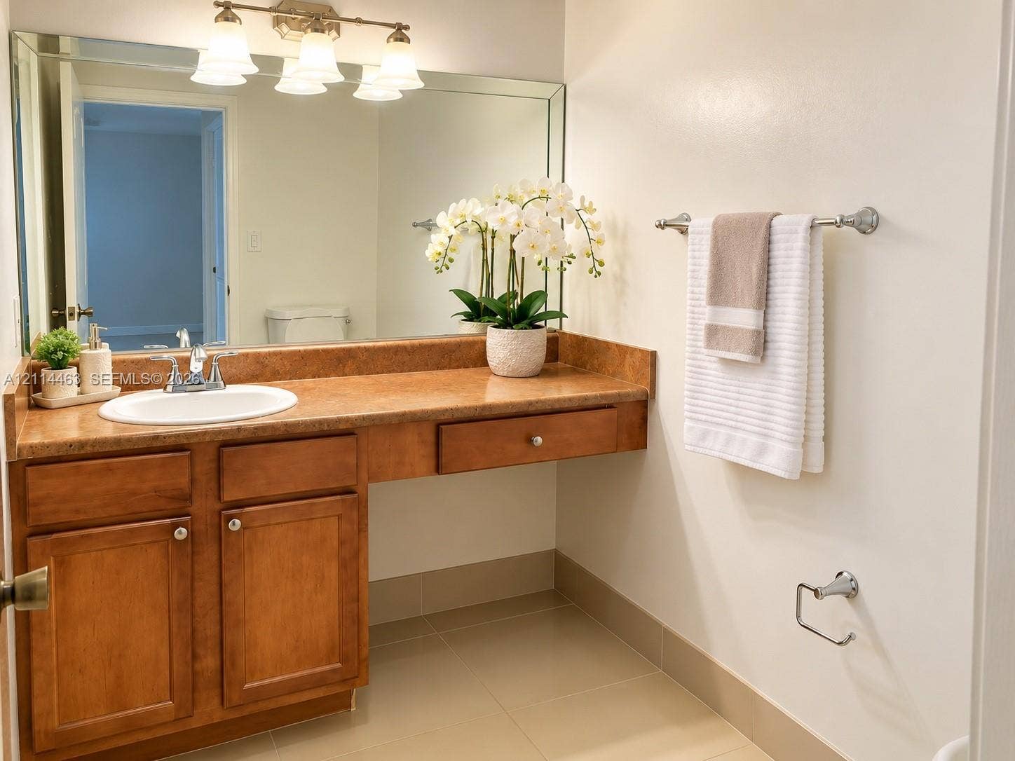 4970 NW 97th Place, #309, Doral, Doral, FL 33178 Bathroom vanity featuring warm-toned cabinetry, an integrated stone-finish countertop with a circular drop-in sink, a large wall-mounted vanity mirror, a three-light fixture, and neutral tile flooring