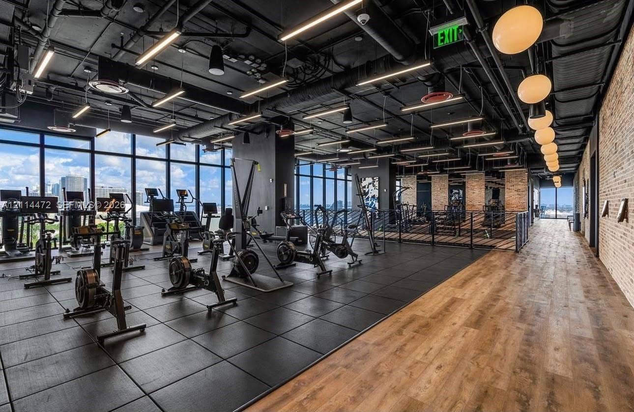 398 NE 5th Street, #4001, Miami, FL 33132 Fitness center featuring floor-to-ceiling windows, exposed industrial ceilings with recessed lighting, wood-finish flooring, and a textured brick accent wall