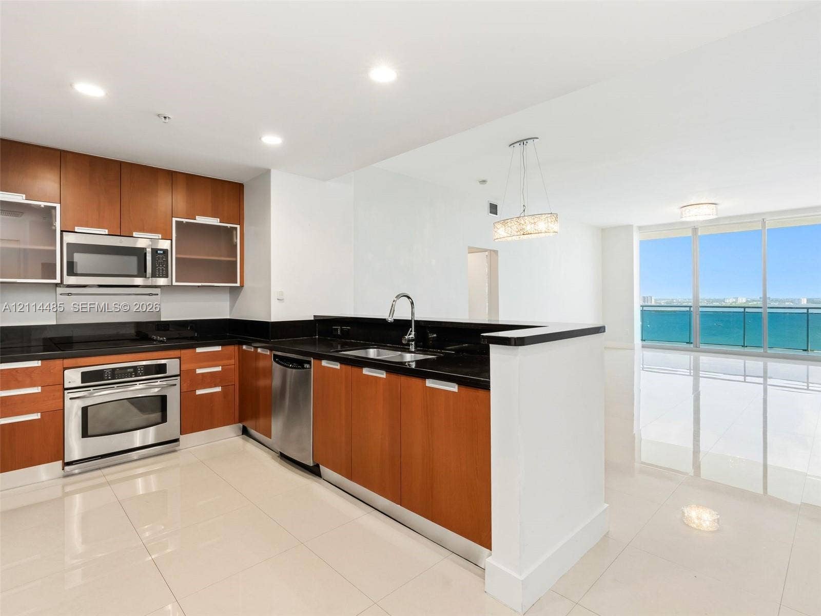 Kitchen featuring wood-finish cabinetry, black countertops, stainless steel appliances, and large-format tile flooring, complete with access to a glass-railed balcony overlooking the water