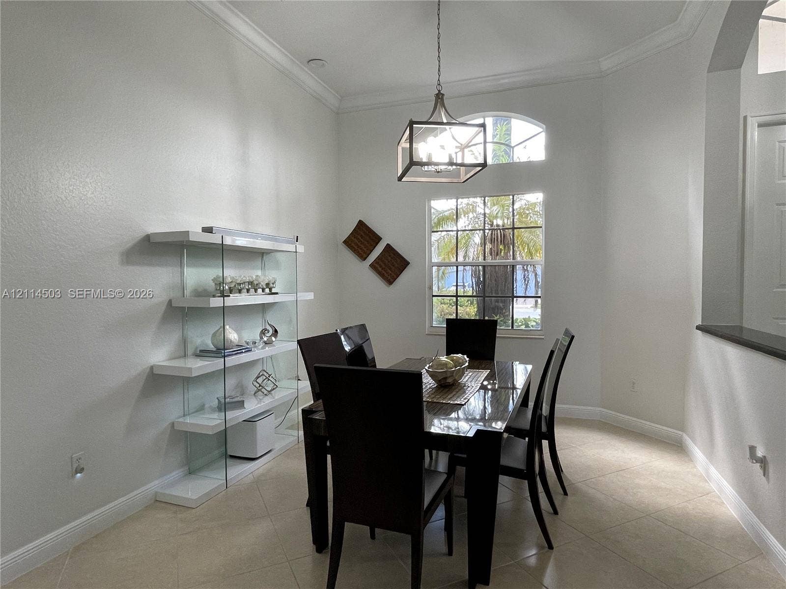 15001 SW 33rd Street, Davie, FL 33331 High-ceiling area featuring crown molding, neutral tile flooring, built-in glass shelving, a multi-light rectangular pendant fixture, and a large window with an arched transom