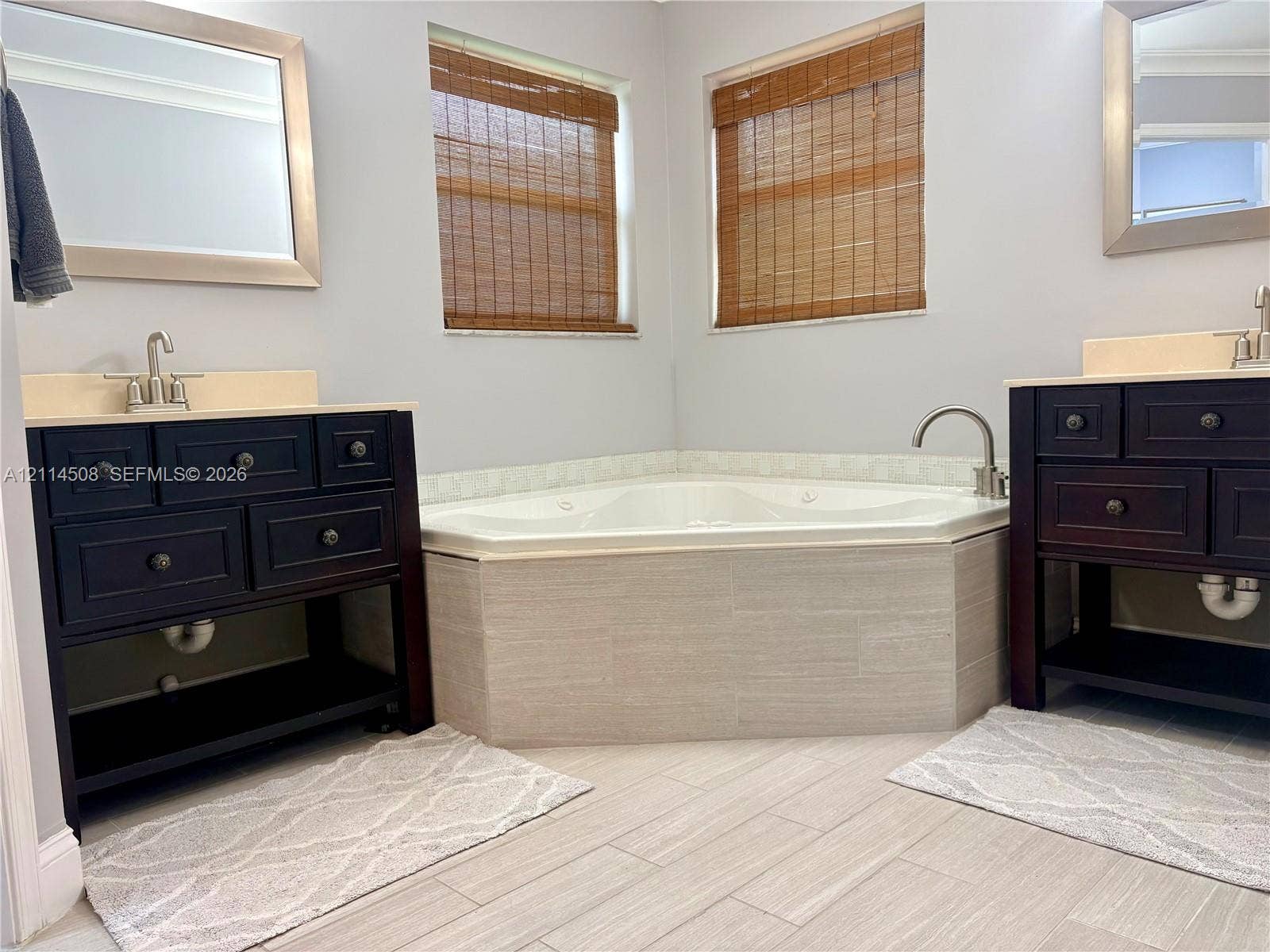 Dual dark-finish wooden vanities with light-toned countertops, corner soaking tub with neutral tile surround, and light-toned tile flooring