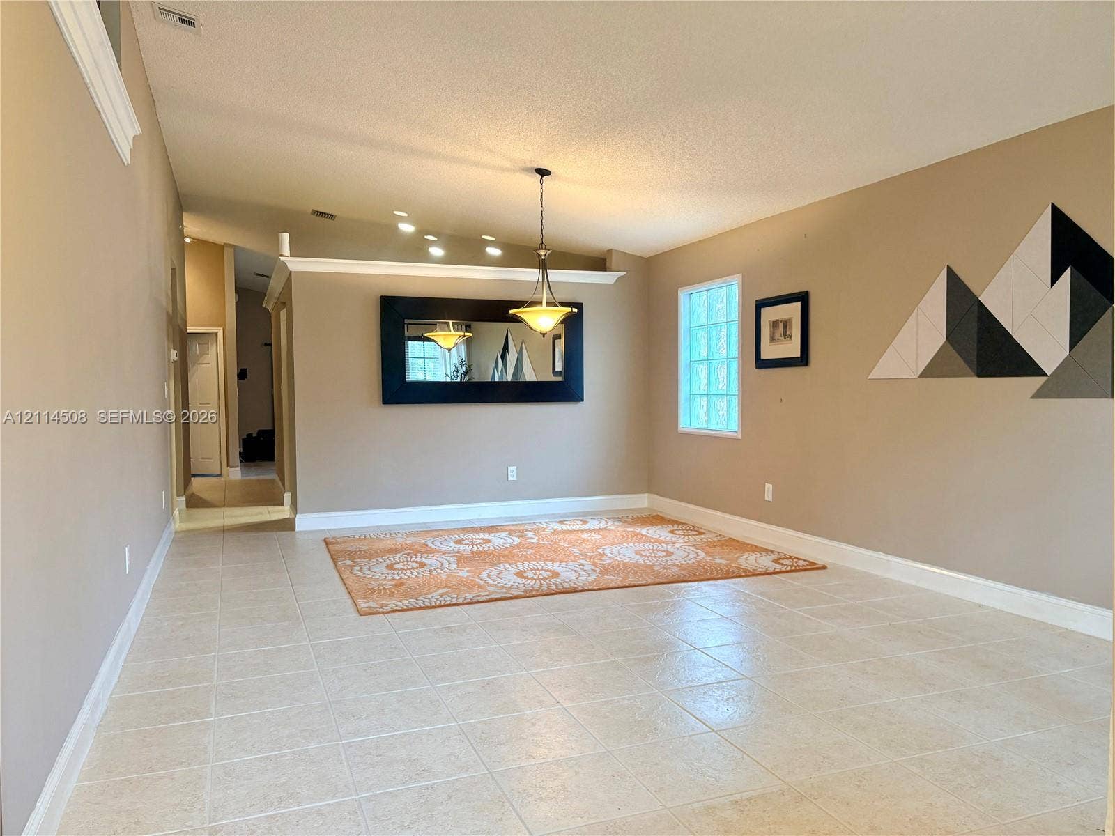 Open-concept interior featuring neutral tile flooring, vaulted ceilings with crown molding, glass block accent window, and a decorative metal pendant light