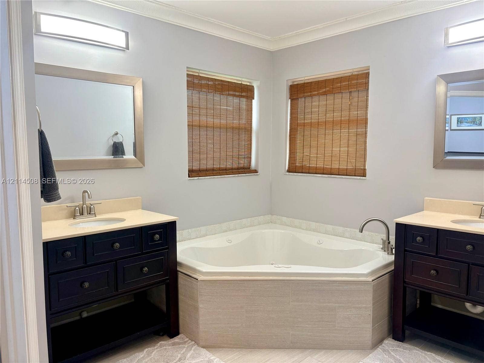 Dual dark wood vanities featuring integrated sinks and stone-surfaced countertops, centered by a built-in corner soaking tub with tile surround and dual wall-mounted light fixtures