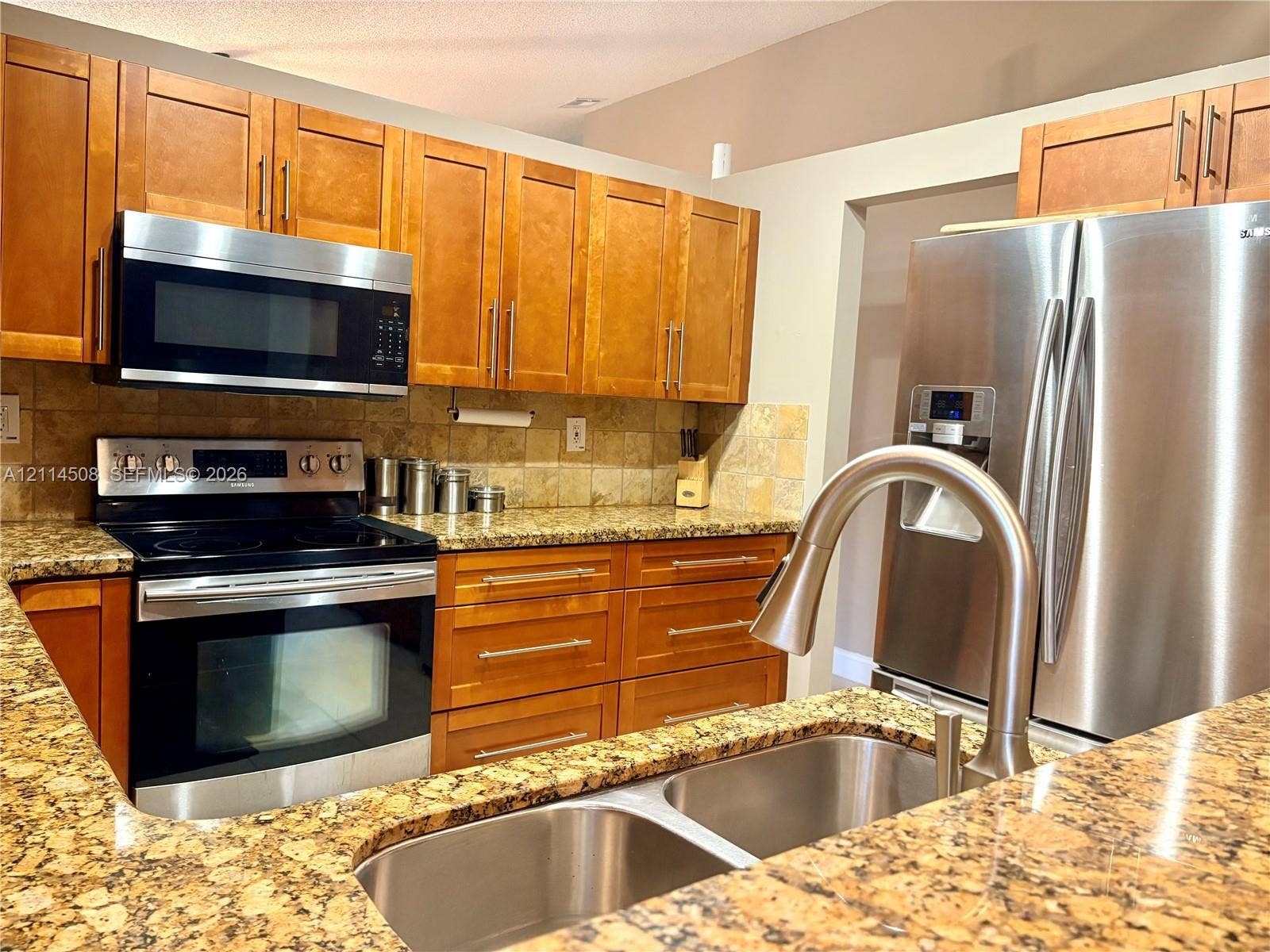 Kitchen featuring natural-finish wood cabinetry, stone-surface countertops, stainless steel appliances, stone tile backsplash, and a dual-basin undermount sink with a high-arc pull-down faucet