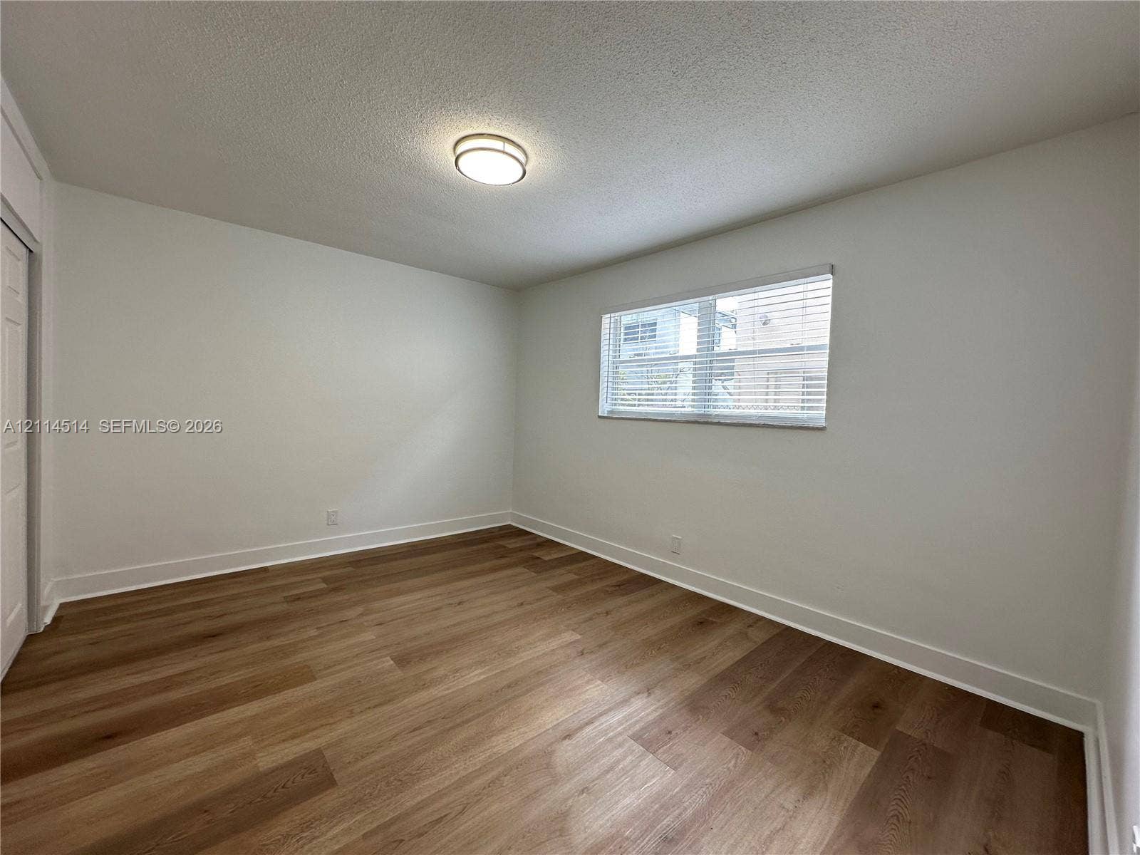 Neutral interior featuring wood-finish flooring, a single-pane window with horizontal blinds, a flush-mount ceiling light fixture, and a standard swinging door