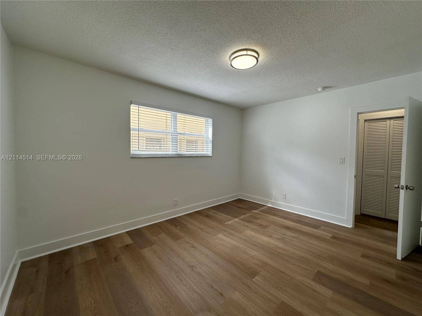 Interior room featuring wood-finish flooring, a neutral color palette, a central flush-mount ceiling light, a horizontal window with slatted blinds, and a closet with louvered double doors