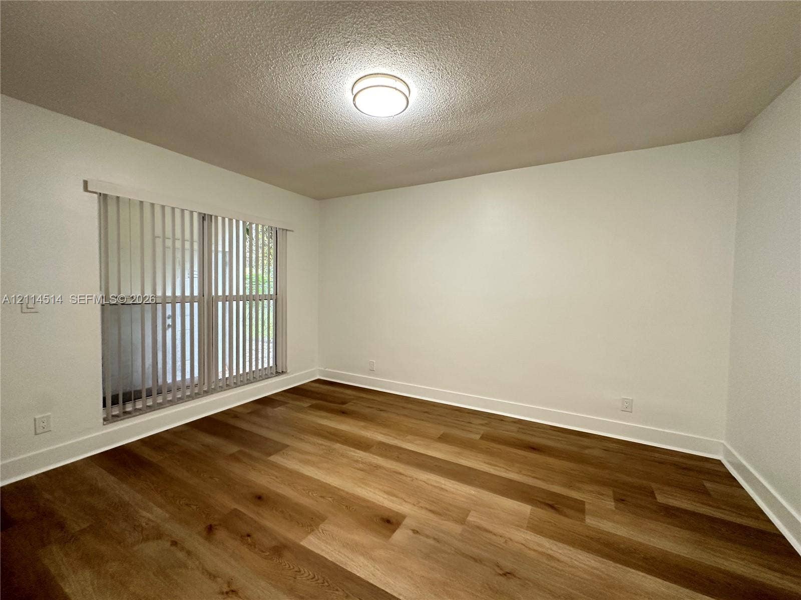 Neutral interior space featuring wood-finish flooring, white walls, vertical blinds, and a central flush-mount ceiling light