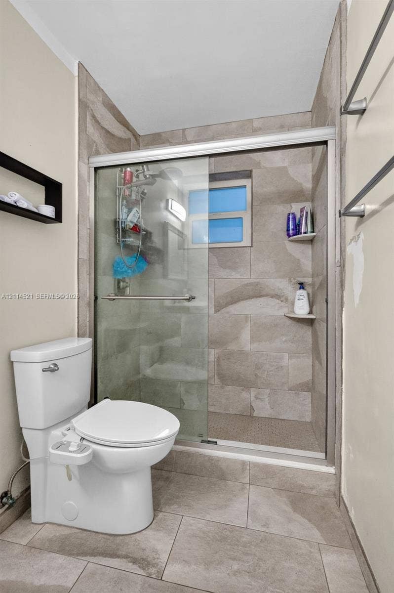 Walk-in shower with sliding glass door, neutral-toned stone-style wall tile, integrated corner shelving, and large-format floor tile