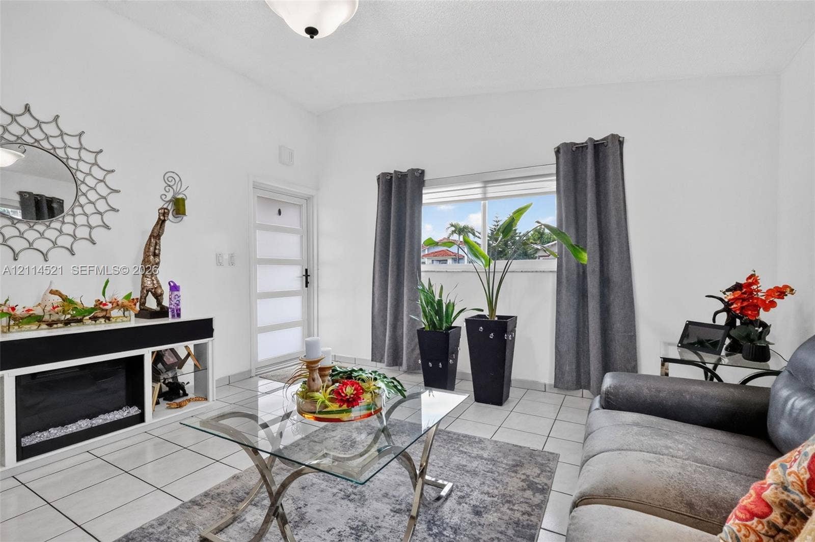 Living space featuring white ceramic tile flooring, a horizontal paneled entry door with frosted glass inserts, a built-in media console, and a large window with blind treatments