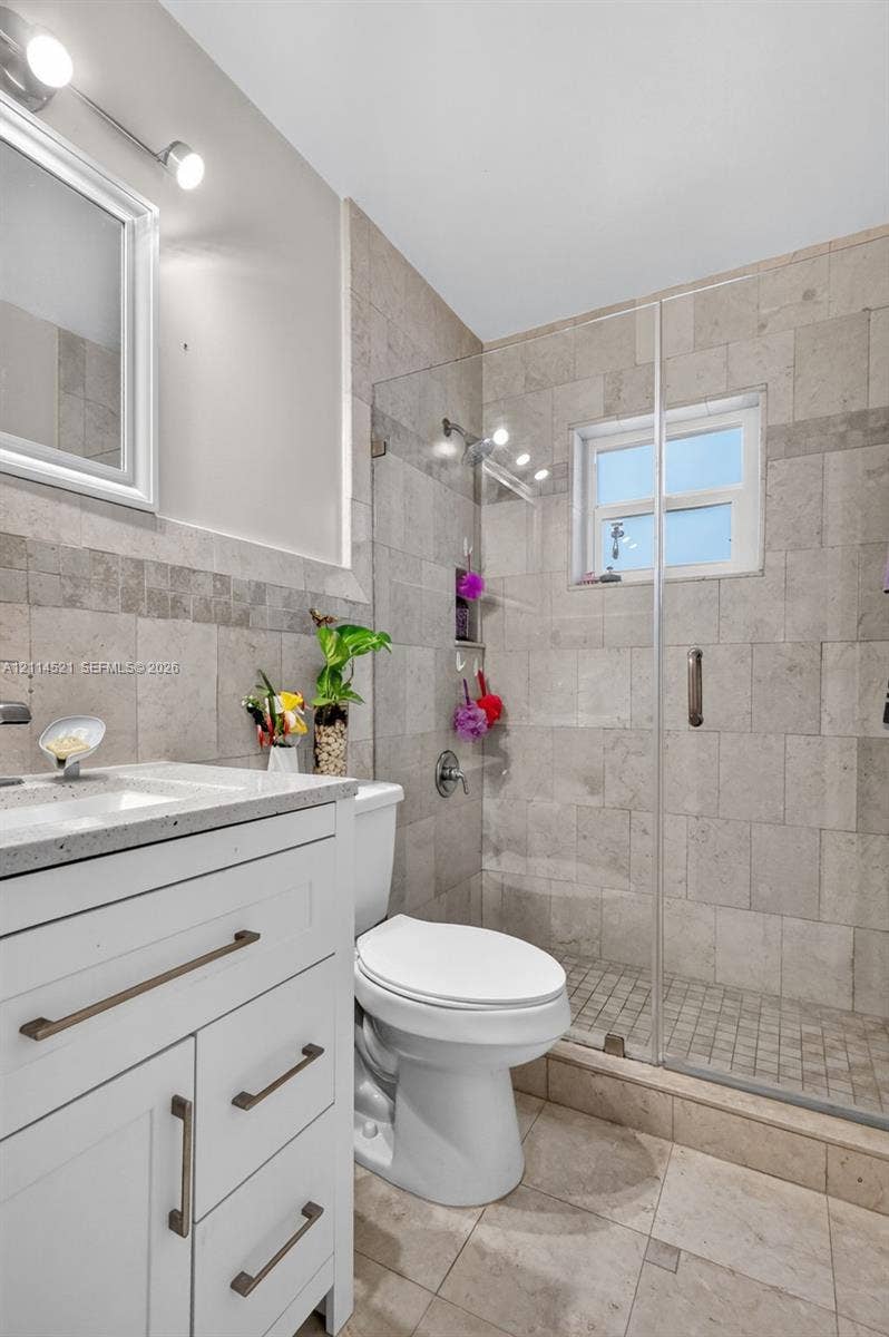 White shaker-style vanity with stone-finish countertop, frameless glass shower enclosure, full-height wall tiling, and dual-light vanity fixture