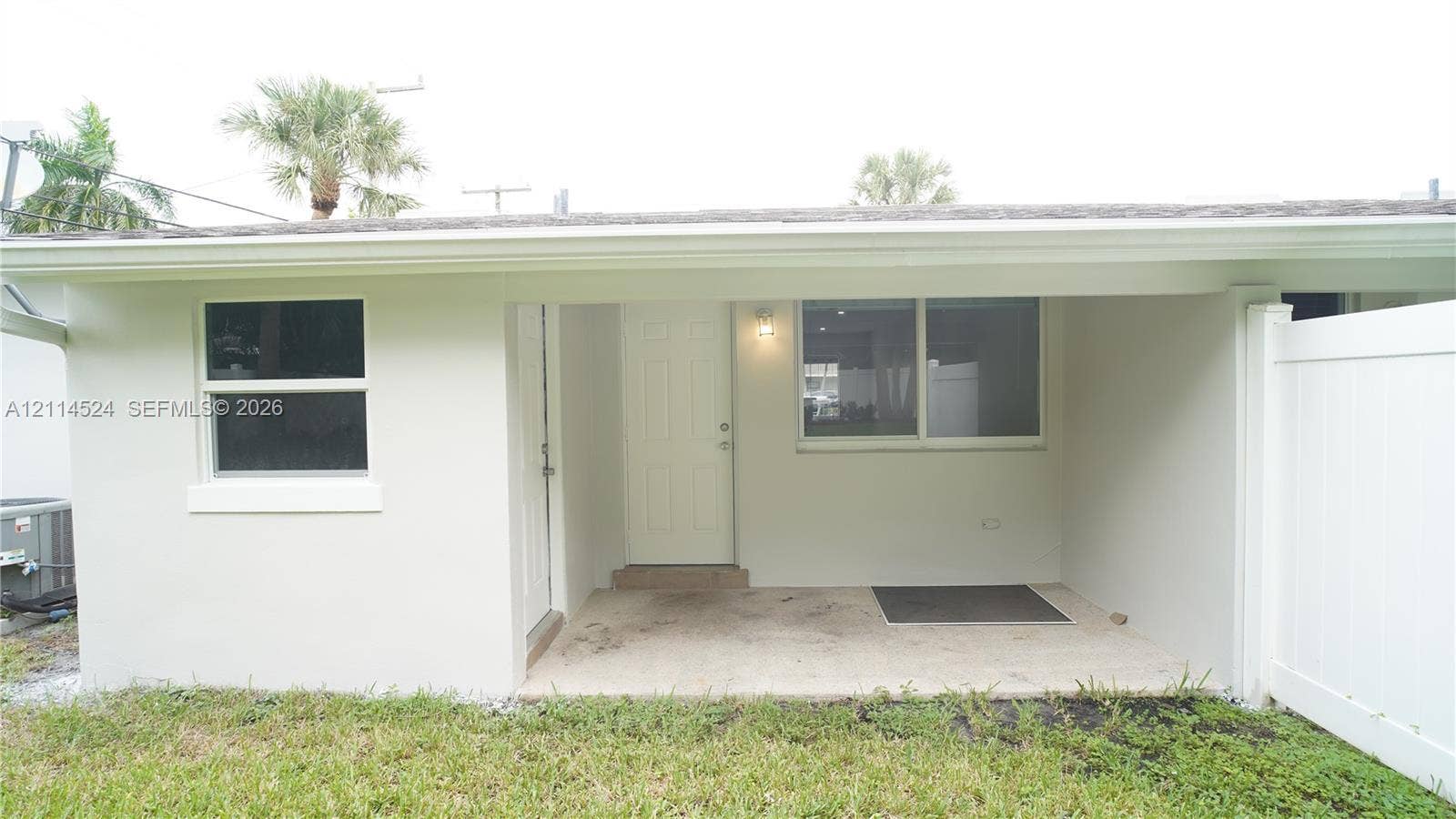 Exterior building facade featuring a covered patio, neutral-toned stucco finish, vinyl-framed windows, and a privacy fence