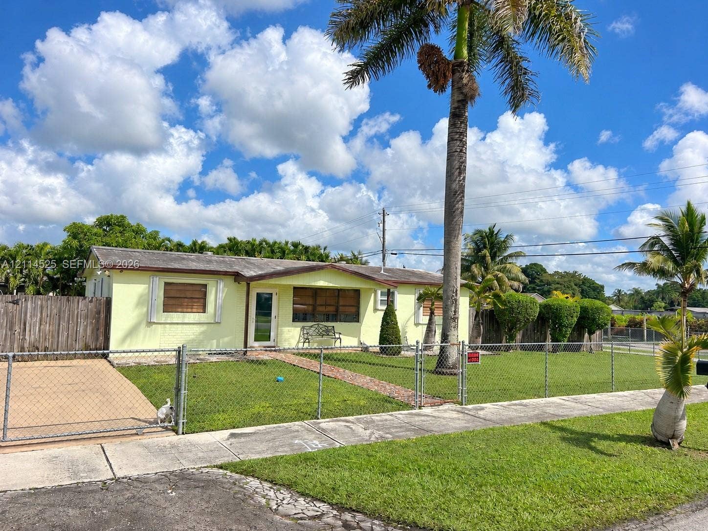 18905 SW 313th Terrace, Homestead, FL 33030 Single-story exterior featuring a brick walkway, chain-link perimeter fencing, mature palm trees, and a paved side driveway