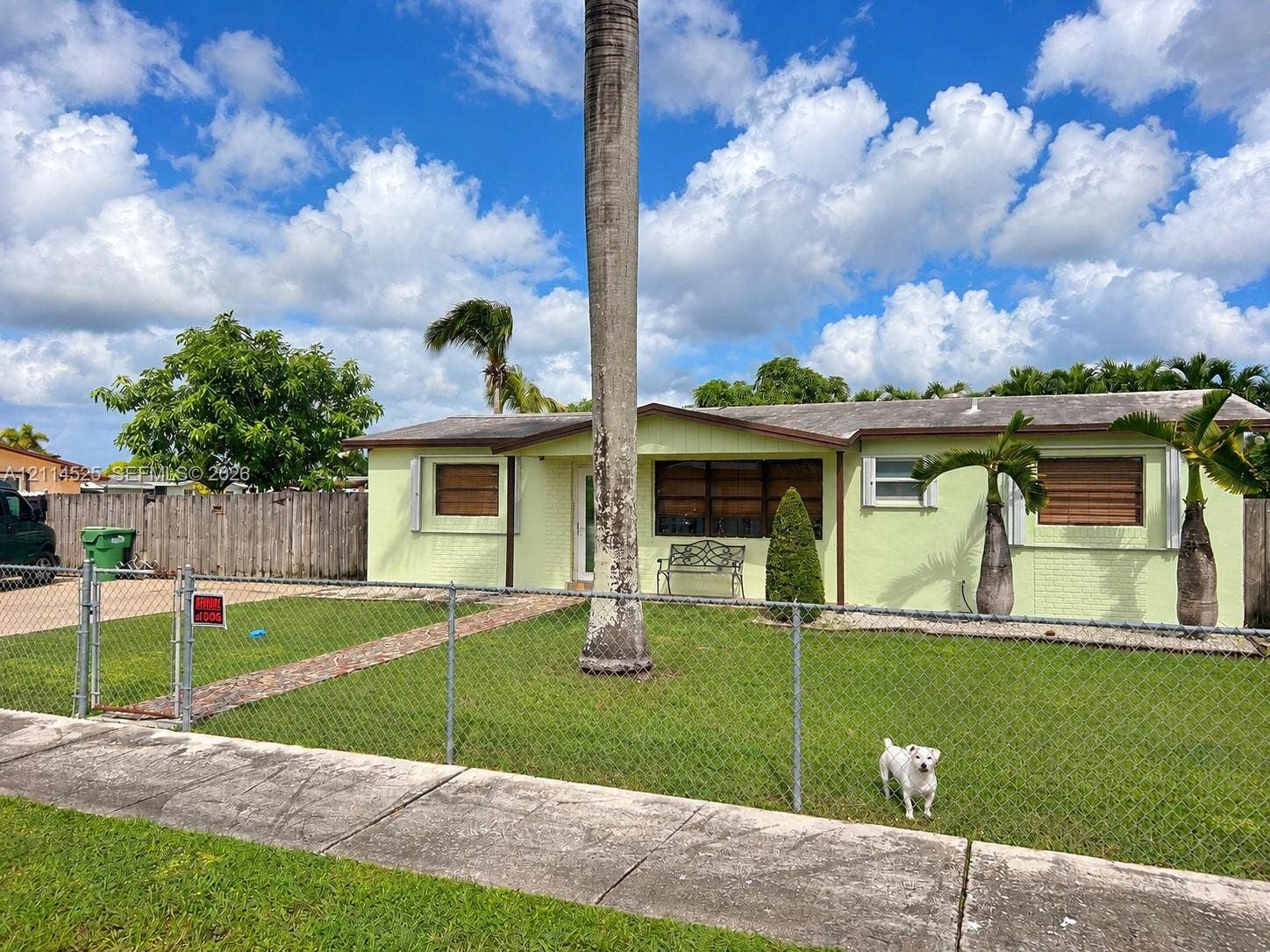 18905 SW 313th Terrace, Homestead, FL 33030 Single-story exterior featuring painted masonry siding, a gabled roofline, metal storm shutters, and a chain-link fenced front yard with mature landscaping