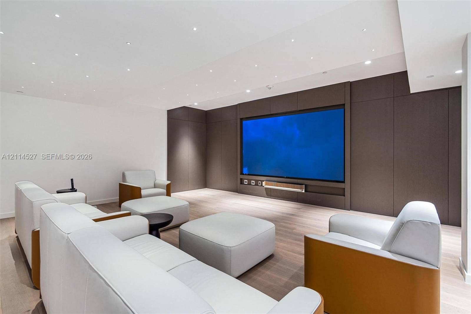 Custom media room featuring a large recessed television screen, built-in floor-to-ceiling paneling, light wood-finish flooring, and recessed ceiling lighting