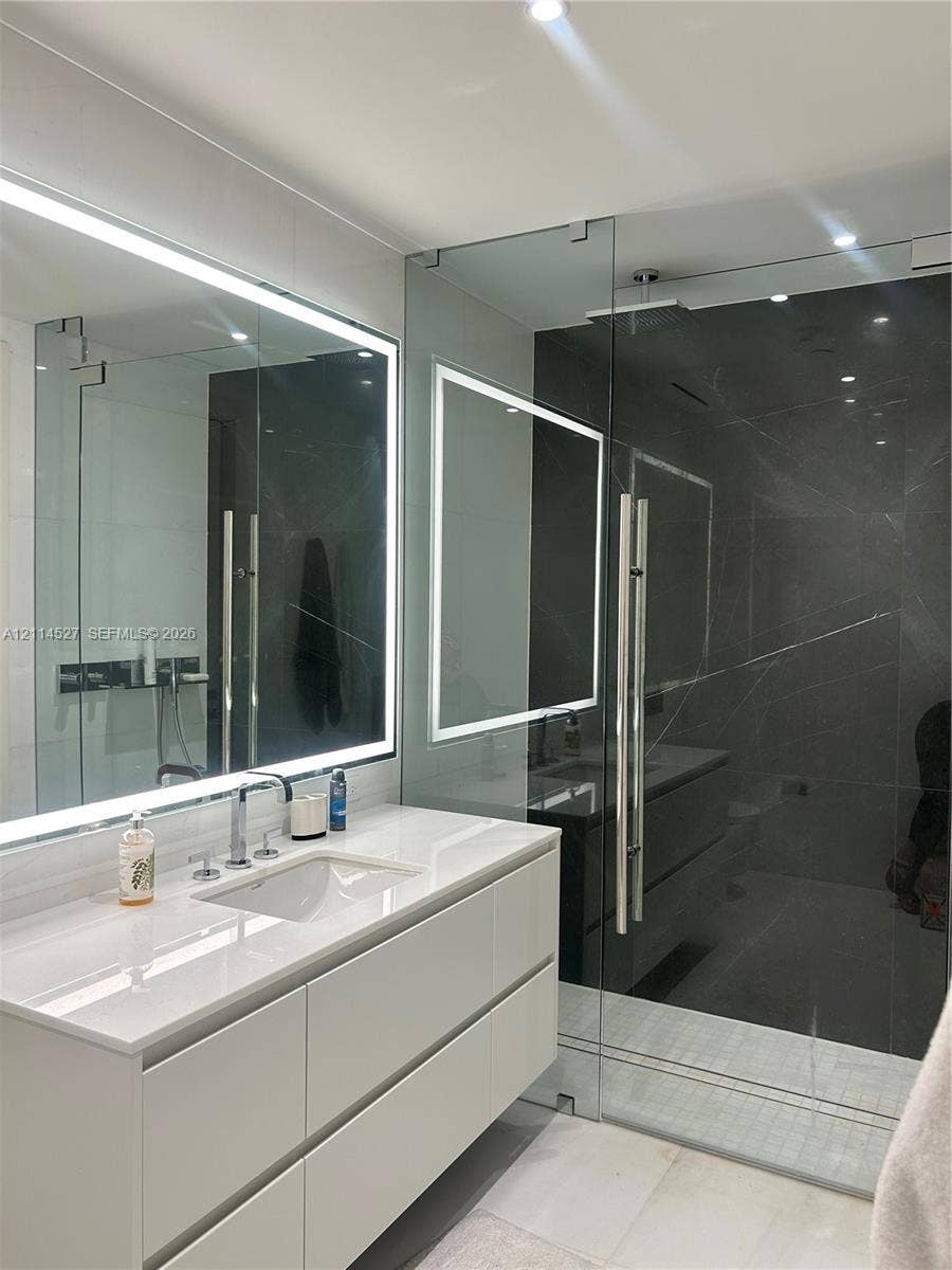 Wall-mounted floating vanity with integrated rectangular sink and stone-surface countertop, frameless glass-enclosed shower featuring large-format dark tile, and integrated LED-backlit mirror