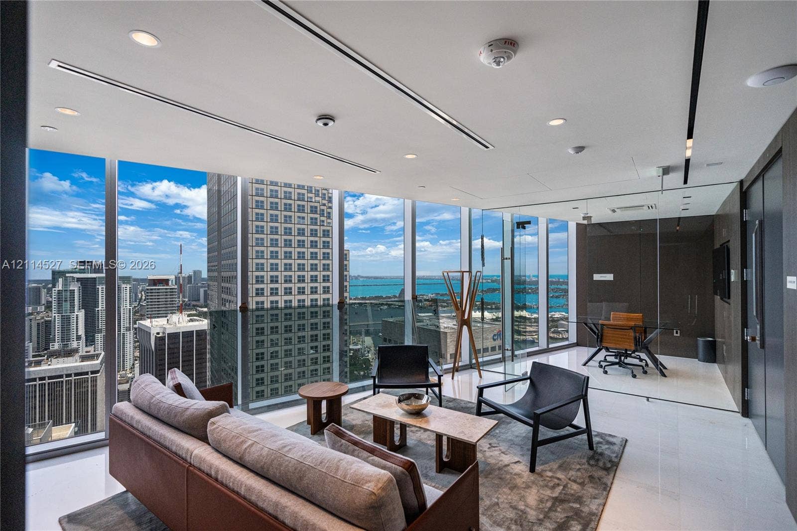 Floor-to-ceiling windows with panoramic cityscape and water backdrops, recessed linear lighting, polished stone flooring, and integrated glass-partitioned office space