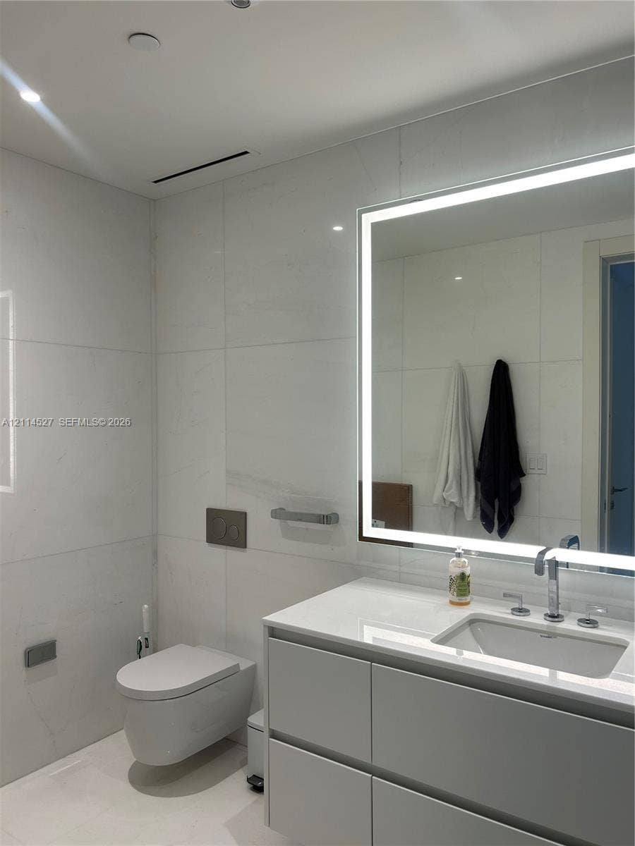 Wall-mounted toilet with a dual-flush actuator plate, large-format tile wall cladding, modern floating vanity with a seamless countertop, rectangular undermount sink, and integrated LED-backlit mirror
