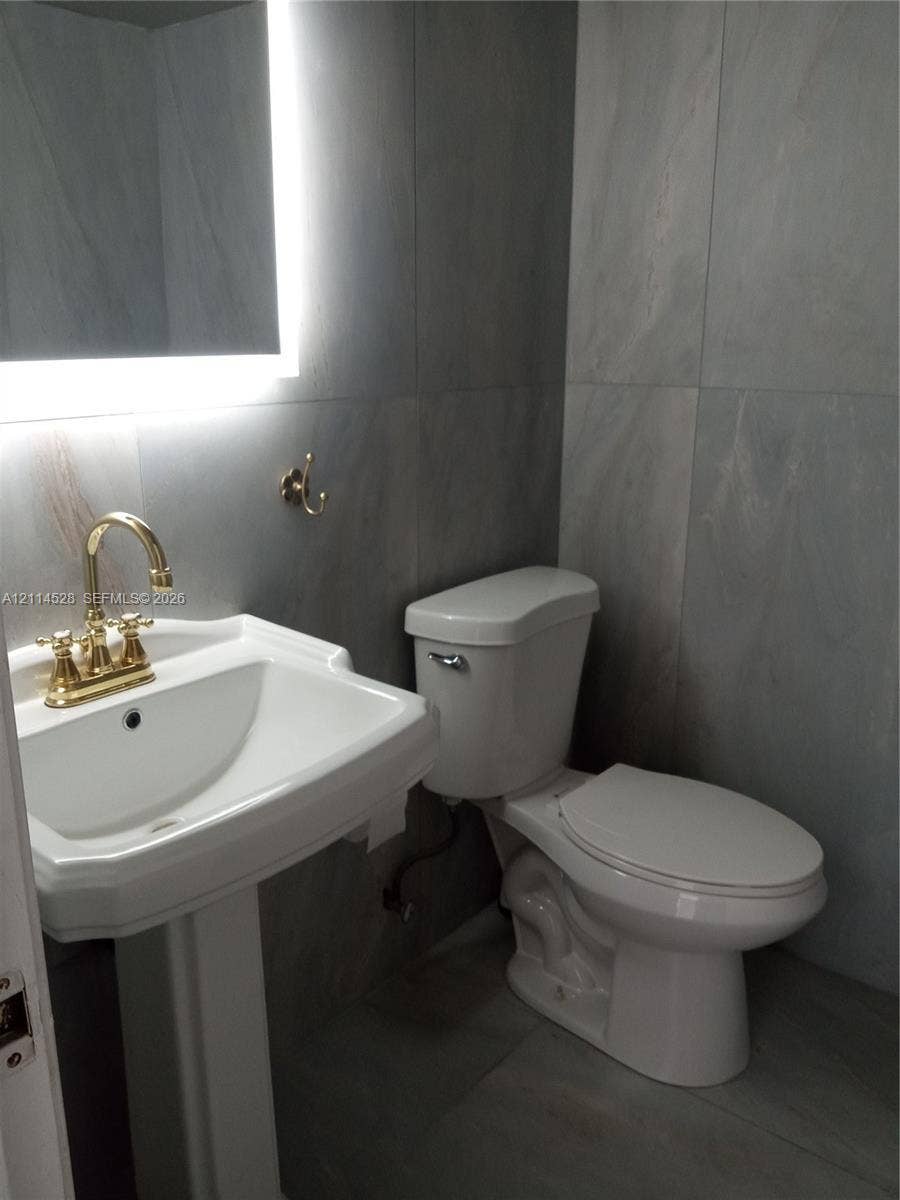 2400 Presidential Way, #1205, West Palm Beach, FL 33401 Pedestal sink with brass finish faucet, large-format grey wall tiling, and backlit vanity mirror