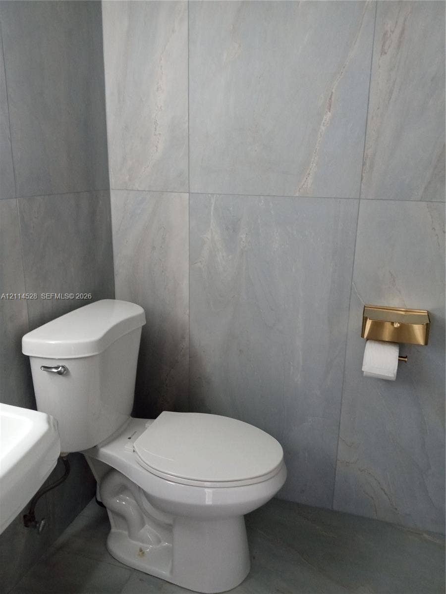 2400 Presidential Way, #1205, West Palm Beach, FL 33401 Full-height large-format wall and floor tiling with subtle veining, dual-flush toilet, and metallic gold-tone toilet paper dispenser