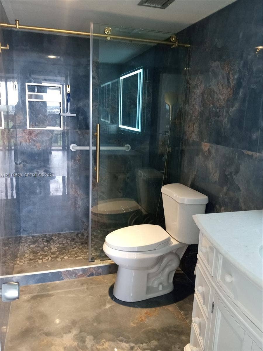 2400 Presidential Way, #1205, West Palm Beach, FL 33401 Walk-in shower featuring gold-tone hardware, a glass sliding door, and a pebble-stone floor, complemented by dark-toned tile walls, a vanity with a light-toned countertop, and a wall-mounted light-up mirror