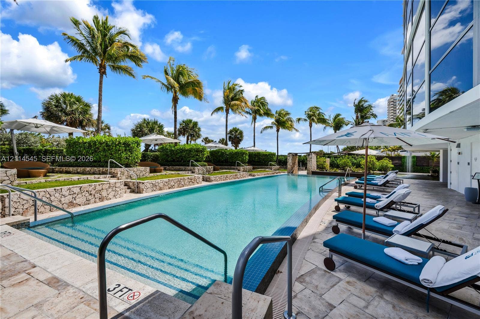5875 Collins Avenue, #1503, Miami Beach, FL 33140 Rectangular swimming pool featuring a tiled waterline, stone-finished decking, tiered landscape planters, and palm tree-lined surroundings