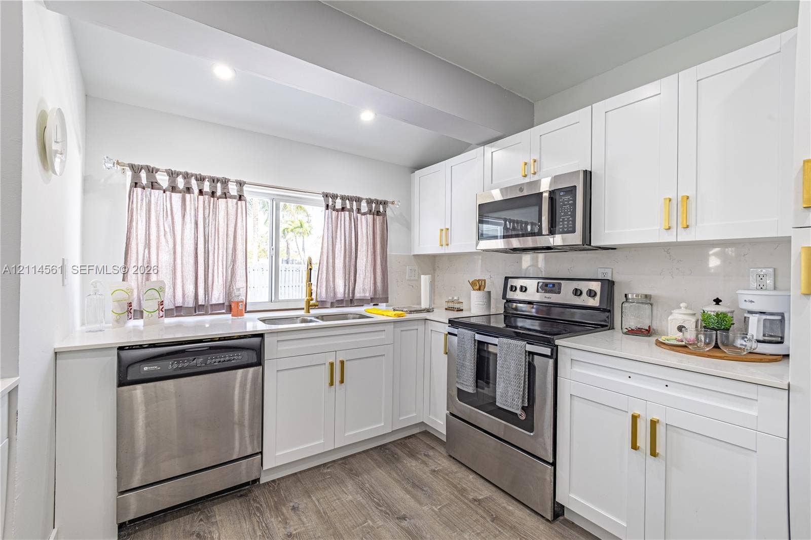 Kitchen featuring white shaker-style cabinetry, metallic gold hardware, stainless steel appliances, light-toned stone-finish countertops, and wood-finish flooring