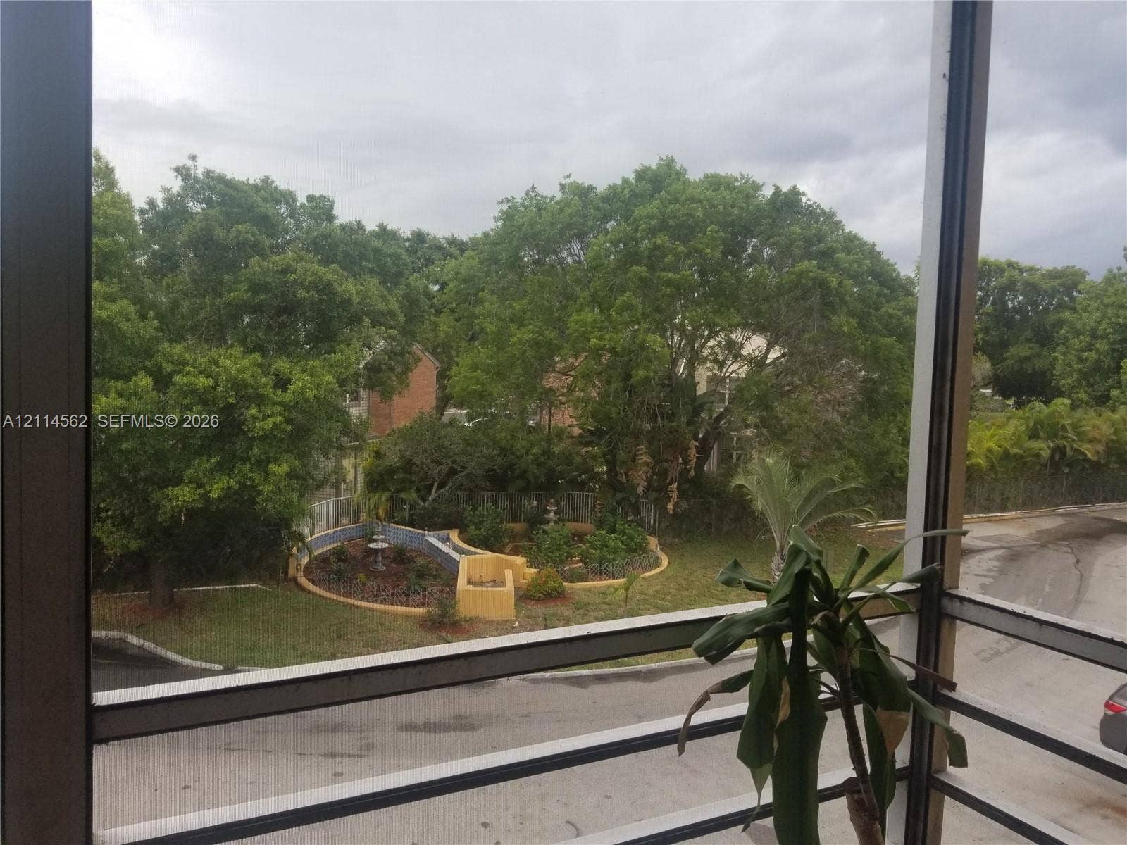 Covered balcony featuring metal railings, overlooking a landscape with mature trees and a masonry garden bed