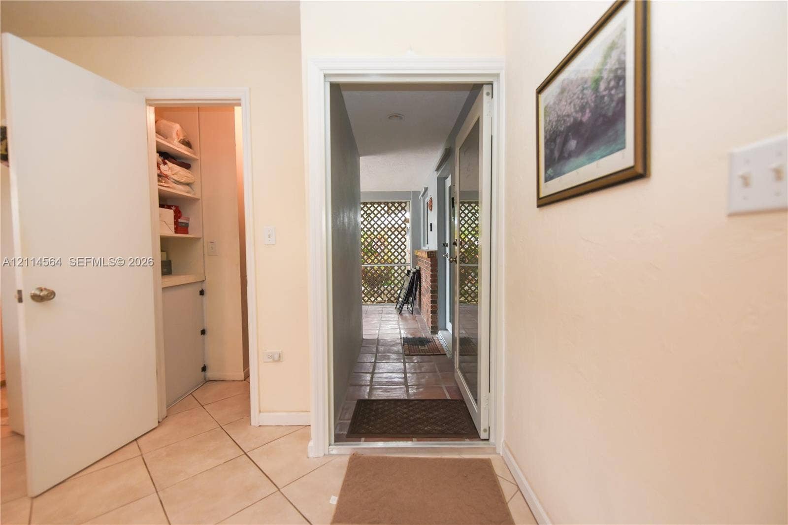 Interior entry featuring diagonal ceramic tile flooring, a built-in reach-in storage closet with multiple shelves, and a glass-paneled exterior door revealing a screened-in porch with brick accents and lattice screening
