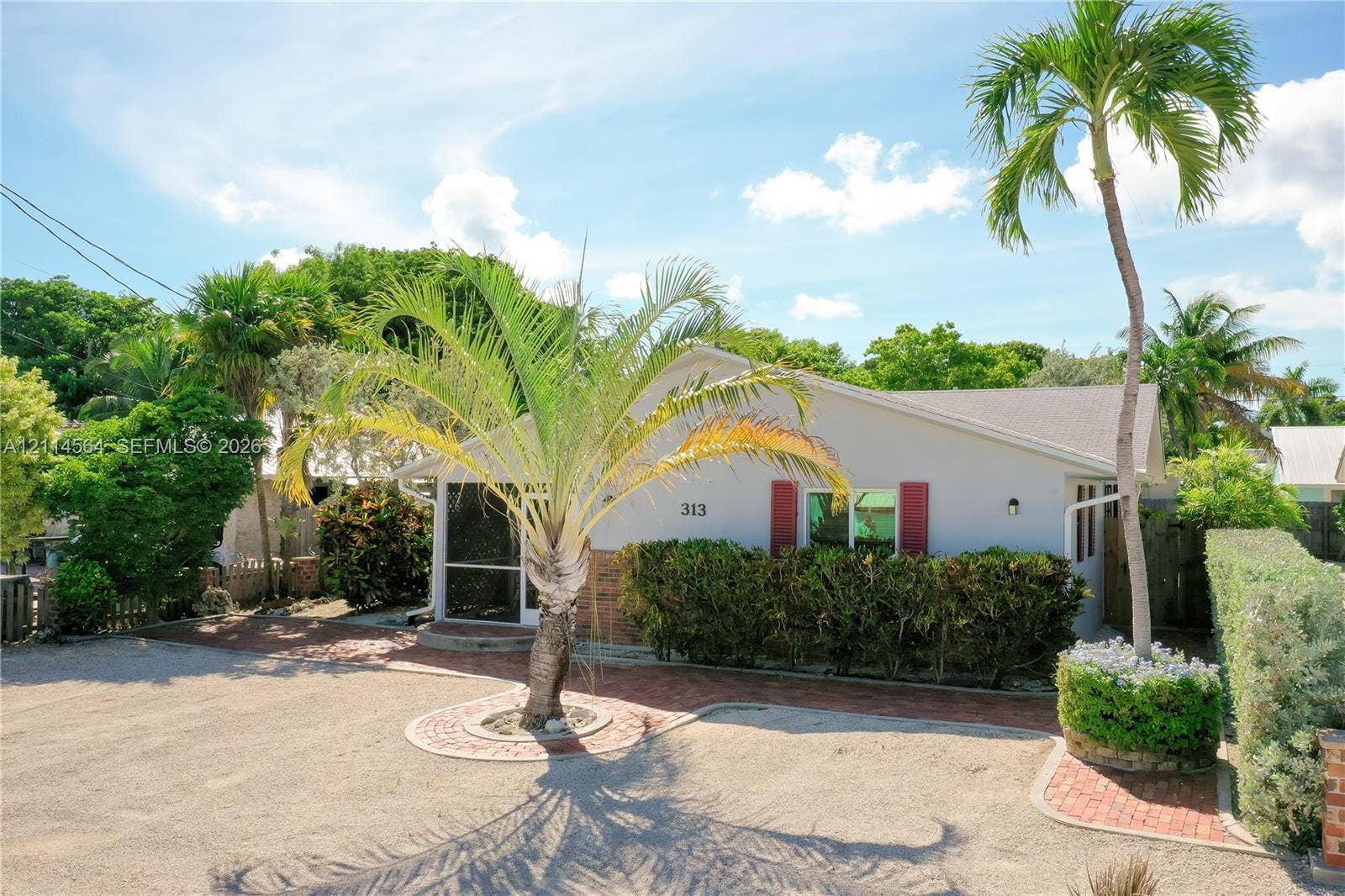 Single-story residence featuring a light-toned exterior, red shutter accents, a screened-in entry, and a gravel parking area with brick-paver borders