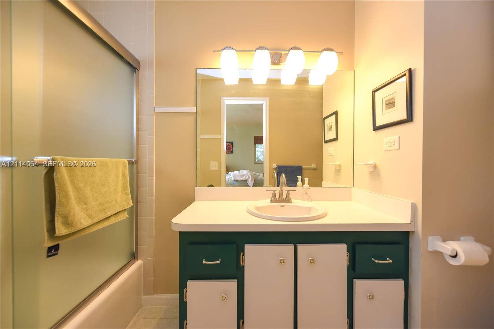 Bathroom vanity featuring a solid-surface countertop, integrated basin, four-light vanity fixture, large wall-mounted mirror, and glass-enclosed shower