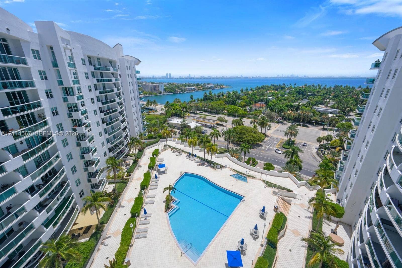 Outdoor swimming pool, paved sundeck, mature palm trees, multi-story building architecture with balconies, and coastal water backdrop