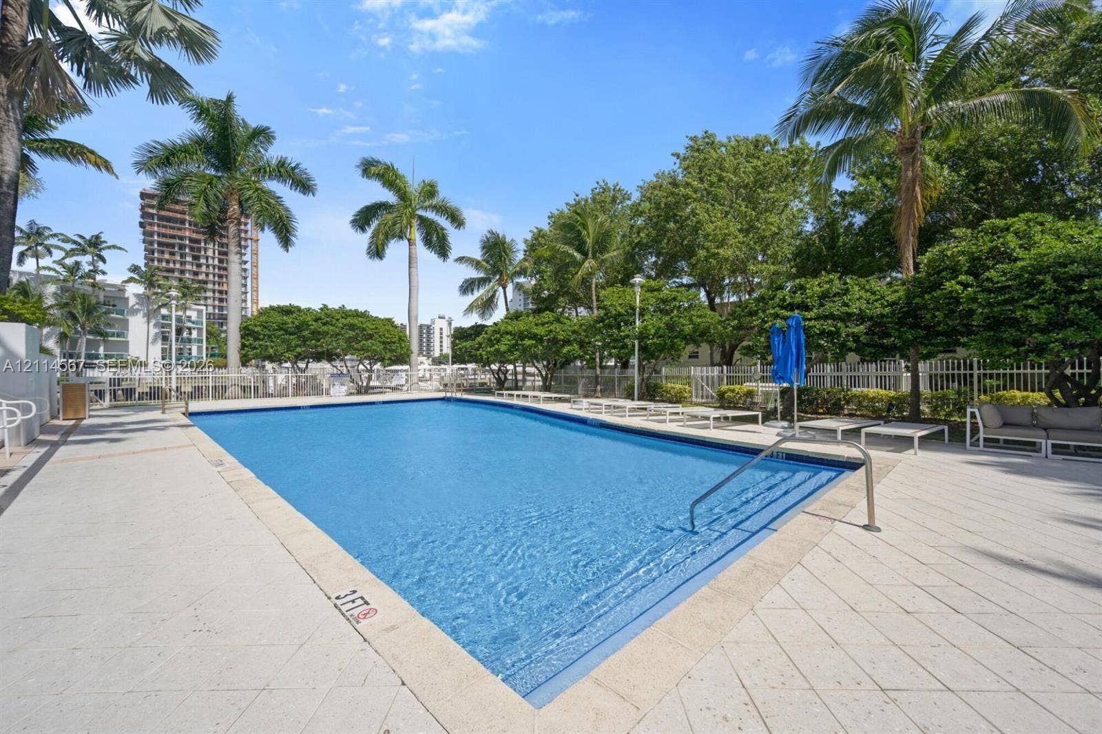 Rectangular swimming pool featuring a tiled deck, metal handrail, palm tree landscaping, and perimeter fencing