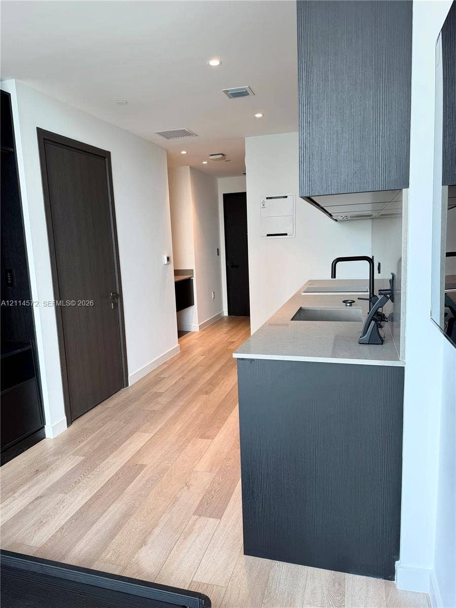 20 NE 11, #3312B, Miami, FL 33132 Kitchenette featuring wood-finish flooring, dark-toned cabinetry, a rectangular stainless steel sink with a matte black faucet, and a quartz countertop