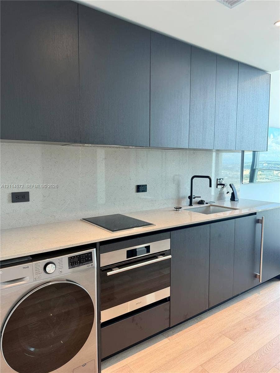20 NE 11, #3312B, Miami, FL 33132 Contemporary kitchen featuring charcoal-toned flat-panel cabinetry, light-toned stone-surface countertops, integrated cooktop, stainless steel wall oven, and light wood-finish flooring