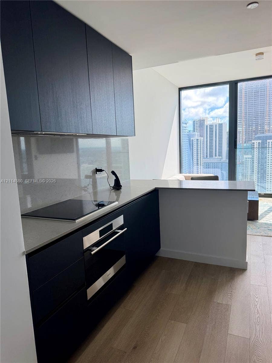 20 NE 11, #4109, Miami, FL 33132 Kitchen featuring dark slab-style cabinetry, solid-surface countertops, integrated cooktop, stainless steel wall oven, and wood-finish flooring
