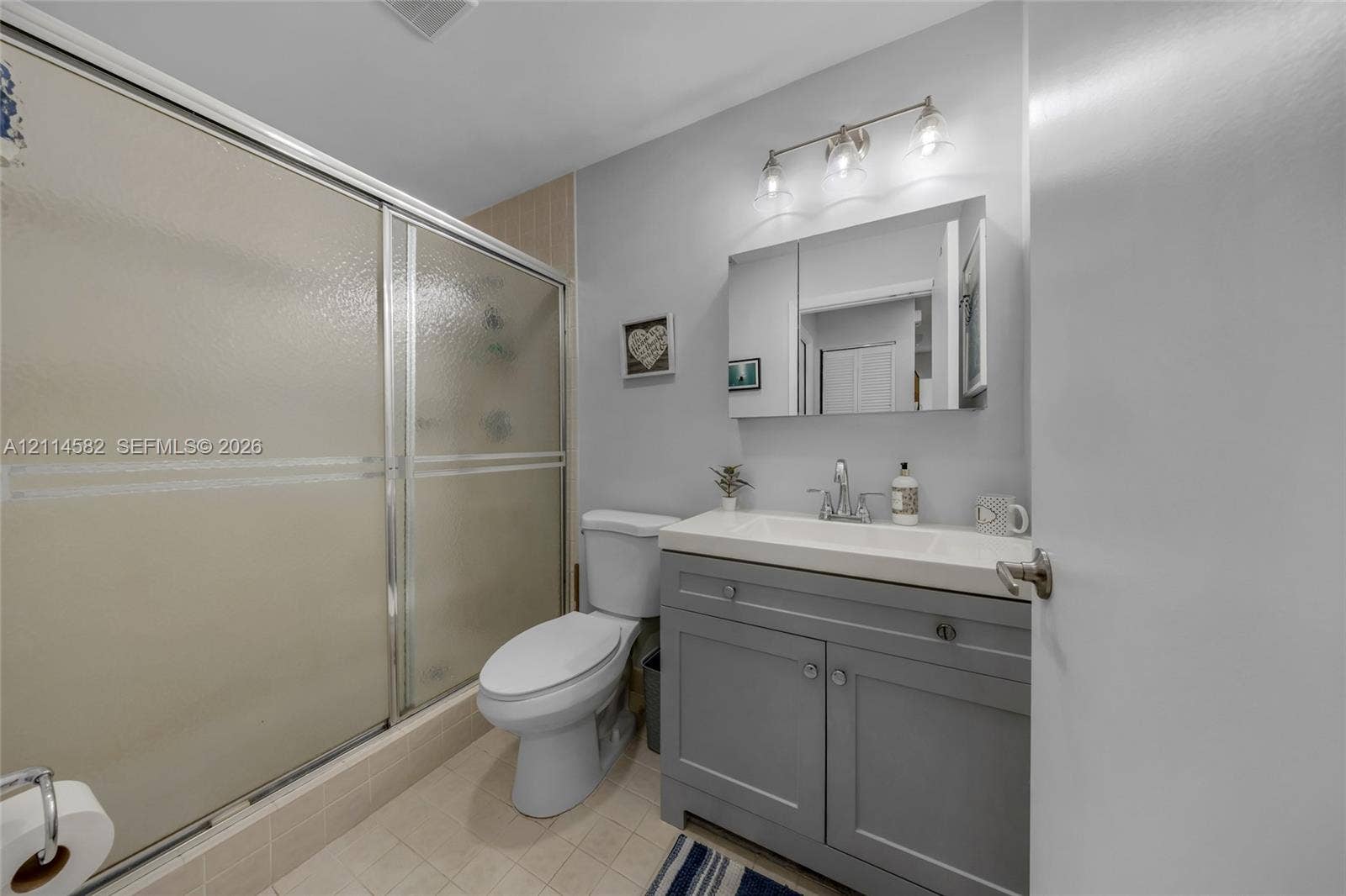 Bathroom featuring a grey shaker-style vanity with an integrated basin, a three-light fixture with glass shades, a mirrored medicine cabinet, and a walk-in shower with a sliding glass door