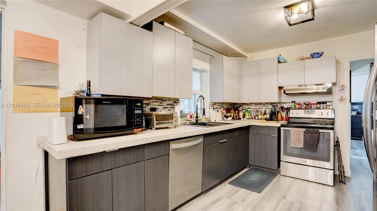 13301 NW Miami Court, Miami, FL 33168 Kitchen featuring two-tone flat-panel cabinetry, light-toned stone-finish countertops, stainless steel appliances, multi-colored tile backsplash, and light-toned wood-finish flooring