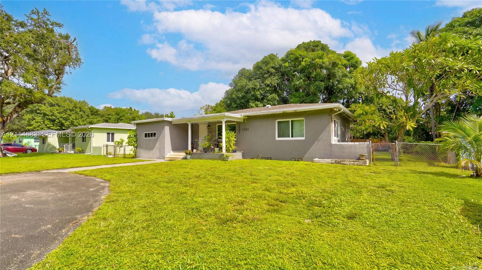 13301 NW Miami Court, Miami, FL 33168 Single-story exterior featuring a gray stucco facade, covered porch entry, asphalt driveway, and expansive lawn with mature landscaping