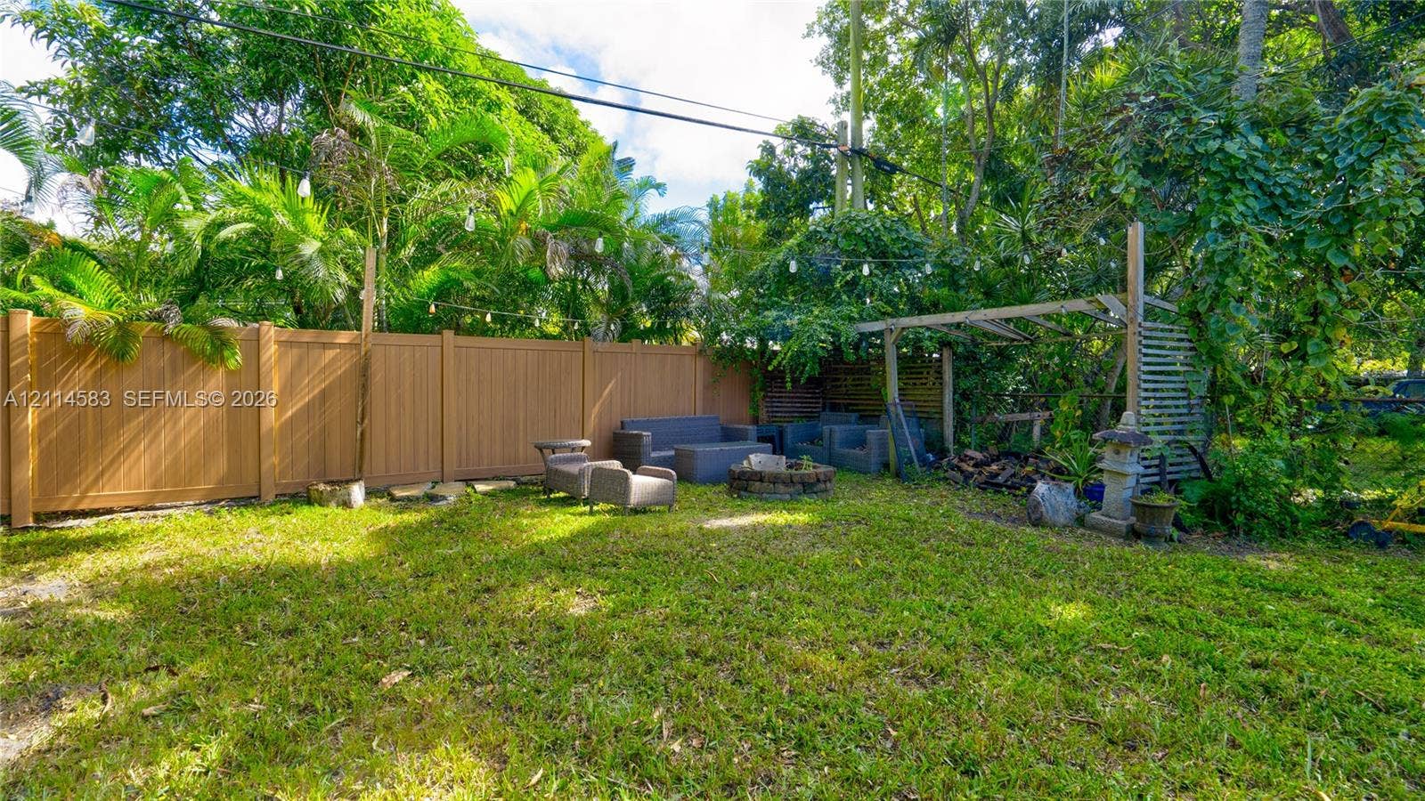 13301 NW Miami Court, Miami, FL 33168 Backyard featuring a privacy fence, stone fire pit, and wooden pergola, surrounded by mature trees and lush greenery