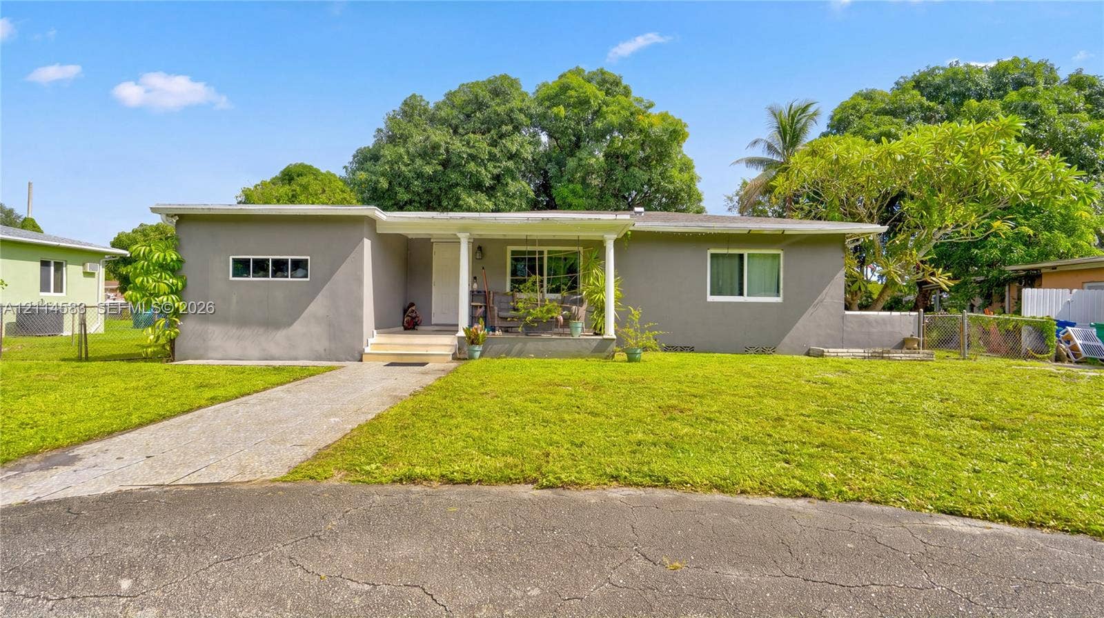 13301 NW Miami Court, Miami, FL 33168 Single-story exterior featuring a gray stucco facade, covered front porch with support columns, concrete driveway, and expansive lawn